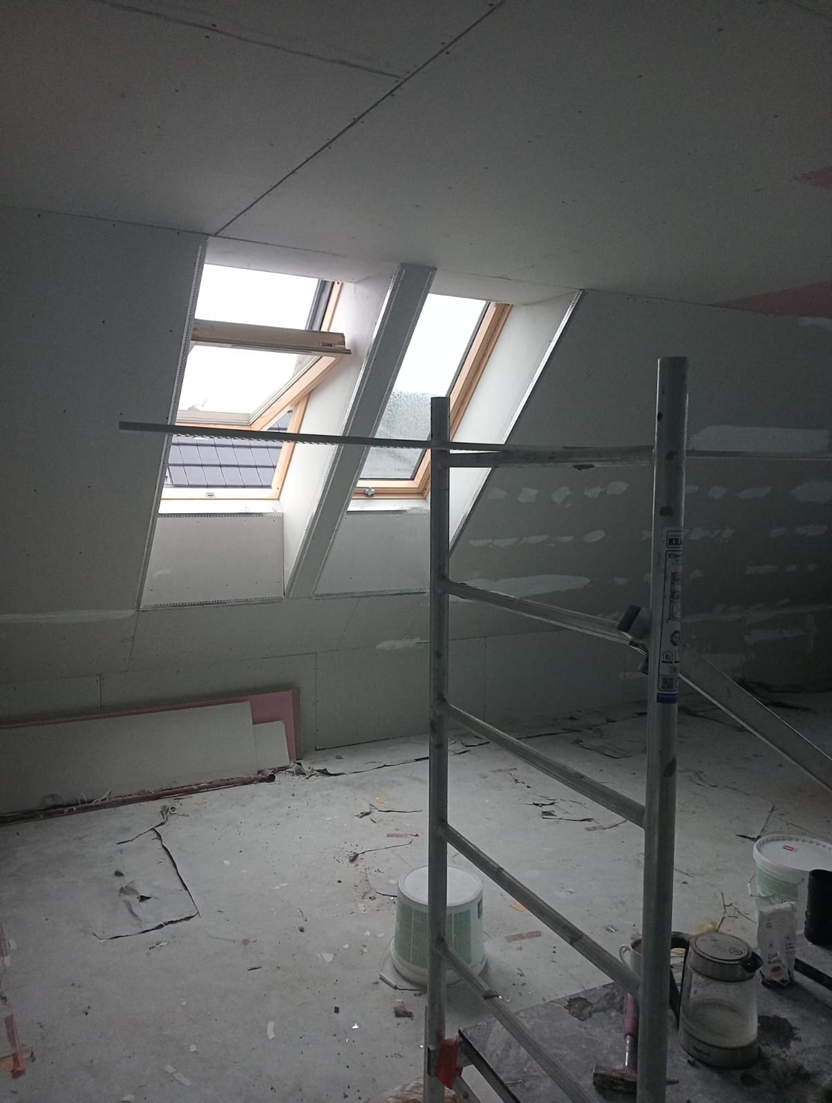 Montaż płyt karton-gips na poddaszu z oknami dachowymi. Widoczne rusztowanie i materiały budowlane. Etap prac wykończeniowych.