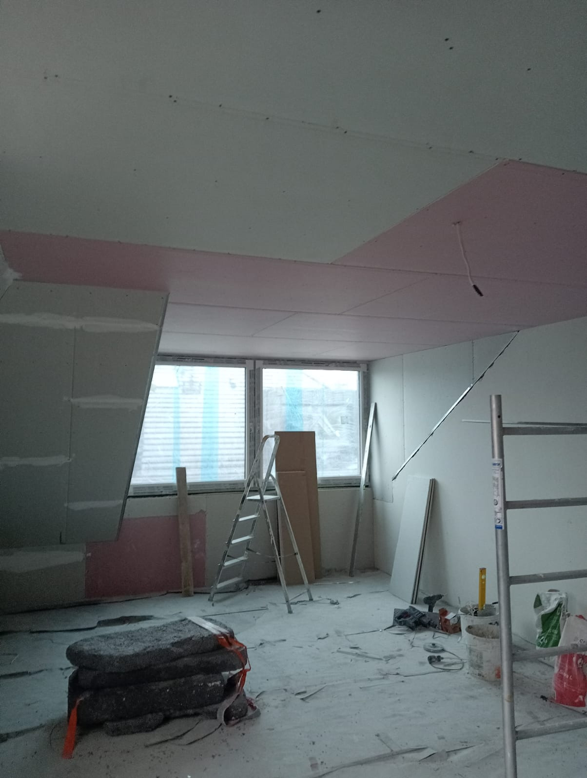 Wnętrze w trakcie montażu płyt karton-gips na suficie, widoczna drabina, narzędzia i materiały budowlane. Częściowo zamontowane płyty z różową warstwą izolacyjną.
