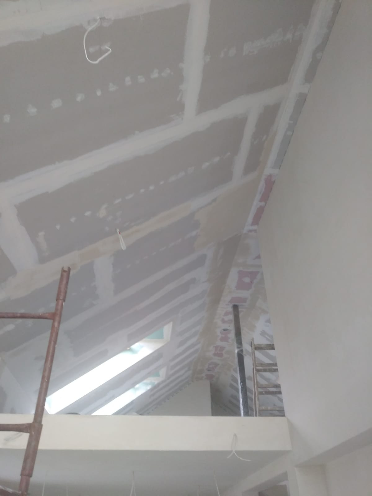 Skośny sufit z płyt karton-gips, na łączeniach widoczne szpachlowanie, w tle okno dachowe i drabina. Prace wykończeniowe na poddaszu.