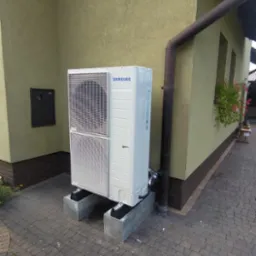 Biała pompa ciepła Samsung na betonowych podstawach, zainstalowana przy ścianie domu z numerem 60 i brązową rynną. Widok z poziomu chodnika z kostki.