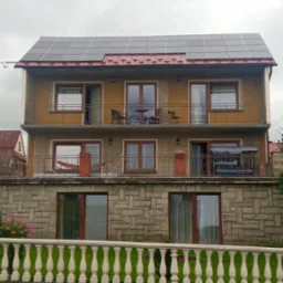 Trzykondygnacyjny dom z balkonami i panelami słonecznymi na dachu, widziany zza ozdobnego ogrodzenia. Widoczne okna PCV i elewacja w odcieniach beżu i szarości.