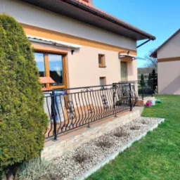 Jasny, jednopiętrowy dom z beżową elewacją, ciemną dachówką i ozdobną, czarną balustradą balkonu. Widoczny fragment ogrodu z zielonym trawnikiem i ozdobnymi kamieniami.