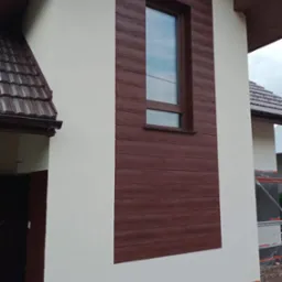 Fragment elewacji domu po malowaniu, z brązowymi panelami imitującymi drewno wokół okna i na dole, oraz jasną, gładką powierzchnią ściany.