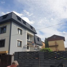 STENO INVESTMENT SPÓŁKA Z OGRANICZONĄ ODPOWIEDZIALNOŚCIĄ - Dwupiętrowy dom z szarym dachem, pracownicy na rusztowaniu przy elewacji. Widoczny fragment ogrodzenia i sąsiedni budynek. Błękitne niebo z chmurami w tle.