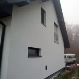 Świeżo otynkowany dom jednorodzinny z szarą stolarką okienną i dachem. Na podw&oacute;rku zaparkowany bus firmy budowlanej. Widok z boku na elewację w pochmurny dzień.