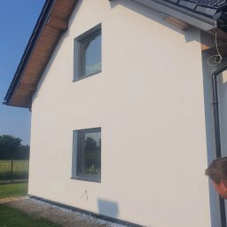 STENO INVESTMENT SPÓŁKA Z OGRANICZONĄ ODPOWIEDZIALNOŚCIĄ - Boczny widok otynkowanego domu jednorodzinnego we Wrocławiu z dwoma oknami i ciemną dachówką. Elewacja w odcieniach bieli. Widoczne fragmenty ogrodu.