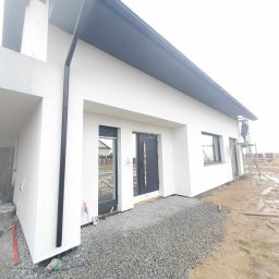 STENO INVESTMENT SPÓŁKA Z OGRANICZONĄ ODPOWIEDZIALNOŚCIĄ - Nowoczesny dom jednorodzinny po malowaniu elewacji, z ciemnoszarymi oknami i dachem. Prace wykończeniowe na zewnątrz, widoczny pracownik na rusztowaniu.