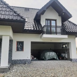 STENO INVESTMENT SPÓŁKA Z OGRANICZONĄ ODPOWIEDZIALNOŚCIĄ - Dom z ciemną dachówką i jasną elewacją, balkon z ozdobną balustradą. Okno zabezpieczone folią, garaż otwarty z przykrytym samochodem. Podłoże żwirowe.