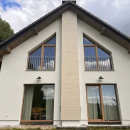 Nowoczesny dom z białą elewacją, drewnianymi oknami i ceglanym kominem, uchwycony w pochmurny dzień z niskiego kąta.