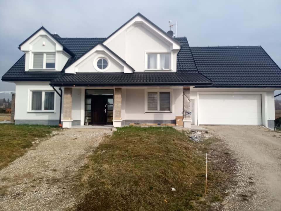 Elewacja domu jednorodzinnego z czarnym dachem i białymi oknami, widok z przodu, z elementami wykończeniowymi przy wejściu i garażu. Ujęcie w pochmurny dzień.