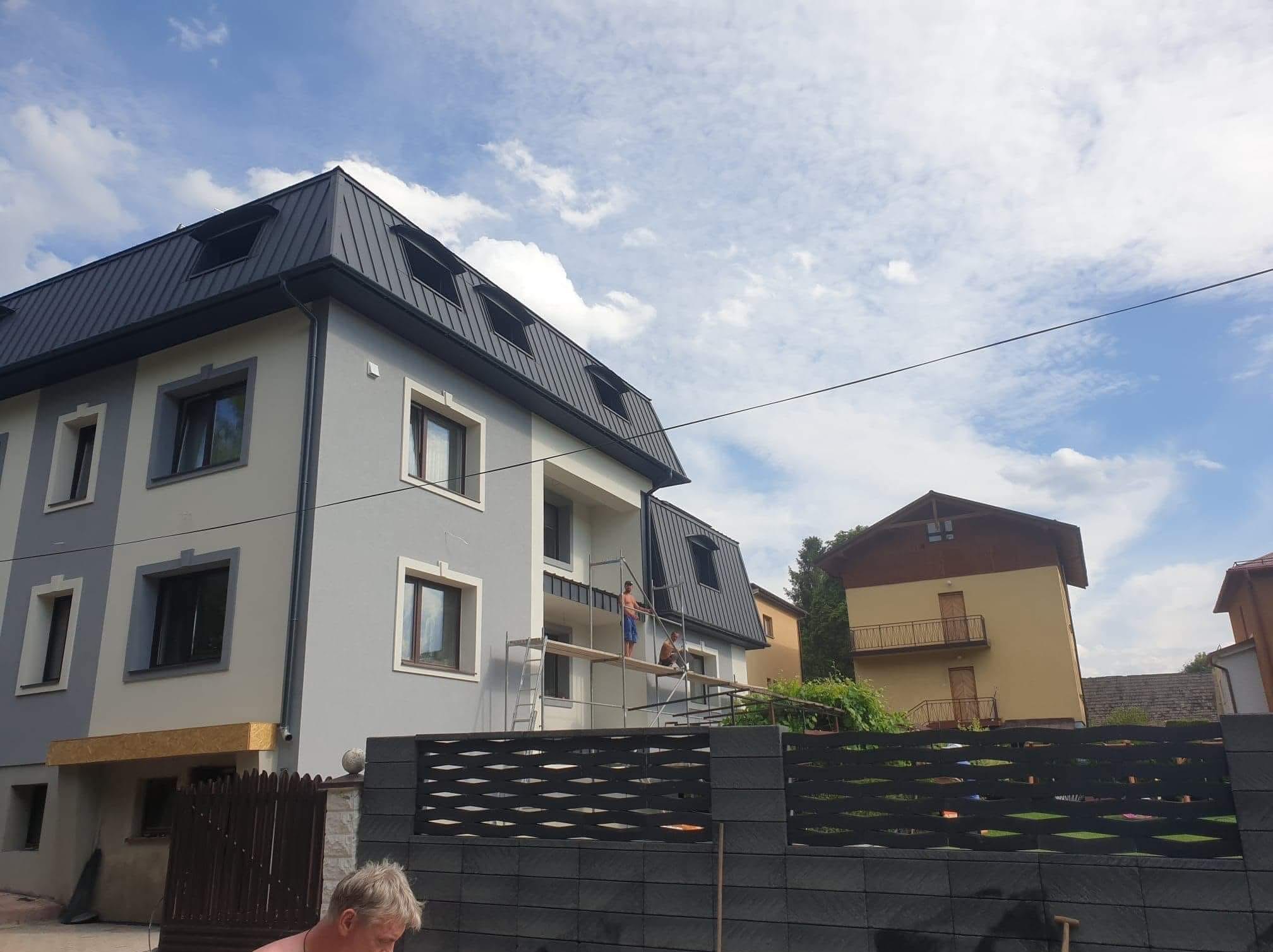Dwupiętrowy dom z szarym dachem, pracownicy na rusztowaniu przy elewacji. Widoczny fragment ogrodzenia i sąsiedni budynek. Błękitne niebo z chmurami w tle.