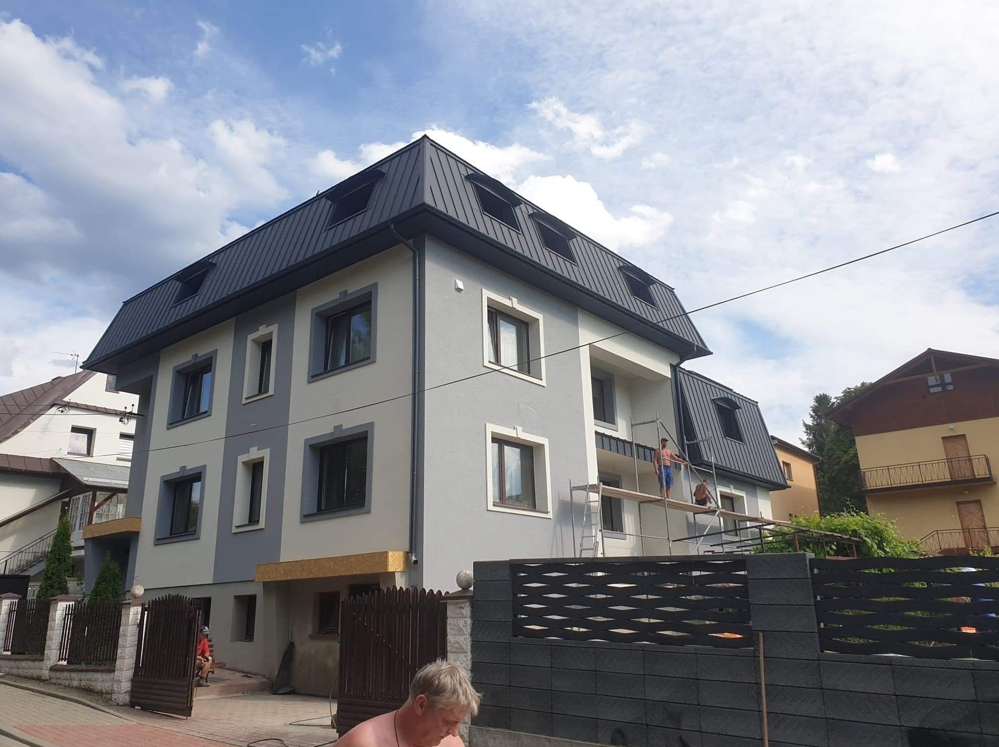 Dwukondygnacyjny dom z szarą elewacją i ciemnym dachem. Na rusztowaniu pracownik. Budynek z oknami w ciemnych ramach i dekoracyjną, ażurową balustradą.