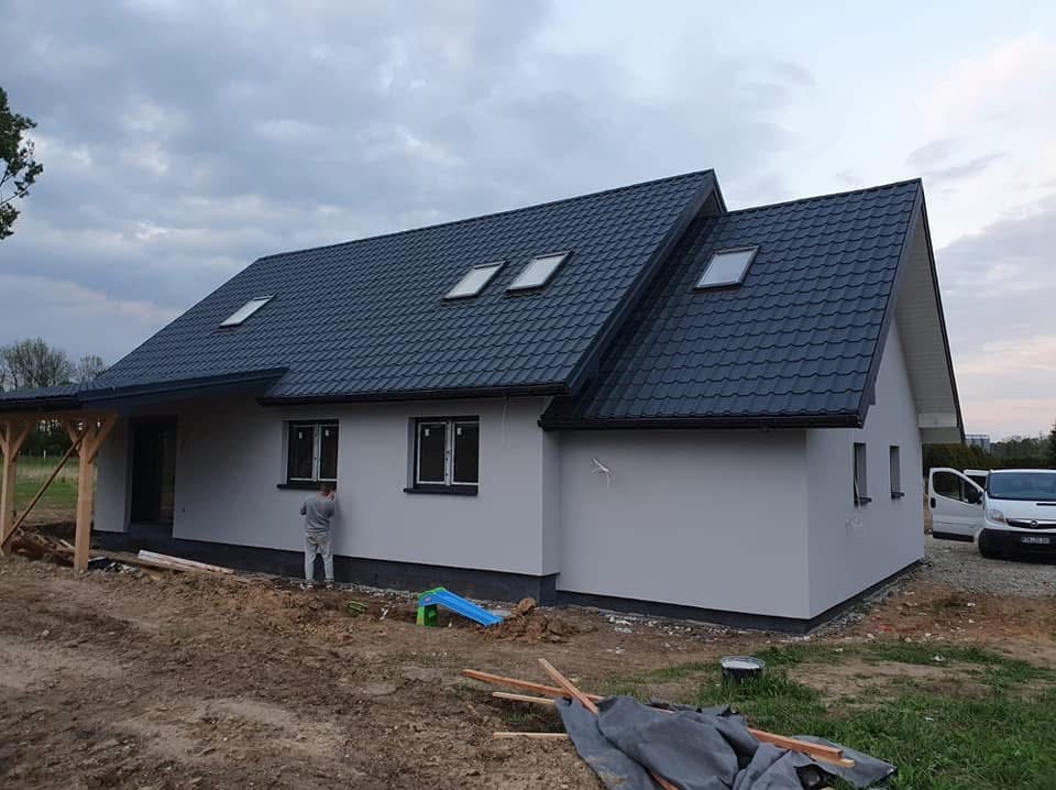 Dom jednorodzinny po malowaniu elewacji na szaro, z ciemnym dachem i oknami dachowymi. Widoczny pracownik przy ścianie, teren wokół domu w trakcie prac wykończeniowych.