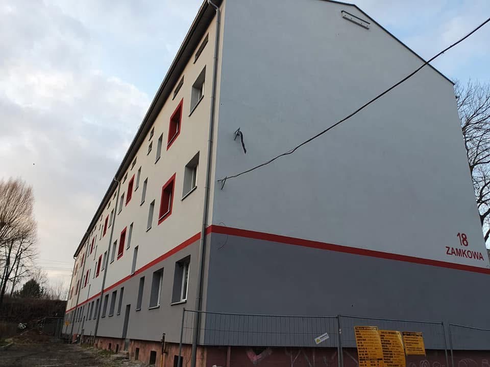 Odnowiona elewacja budynku wielorodzinnego z szarą dolną partią, białymi ścianami i czerwonymi obramowaniami okien. Widoczny numer budynku '18 ZAMKOWA'.