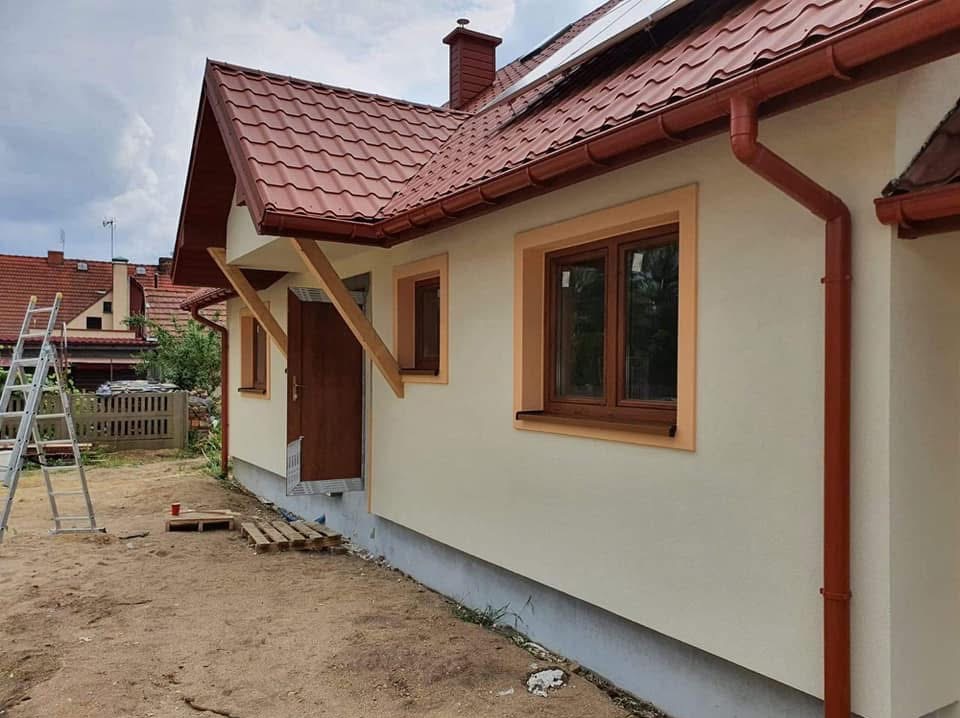 Wykończona elewacja domu z nową stolarką okienną i brązowymi rynnami. Widoczne drewniane podpory nad drzwiami wejściowymi i drabina w tle.