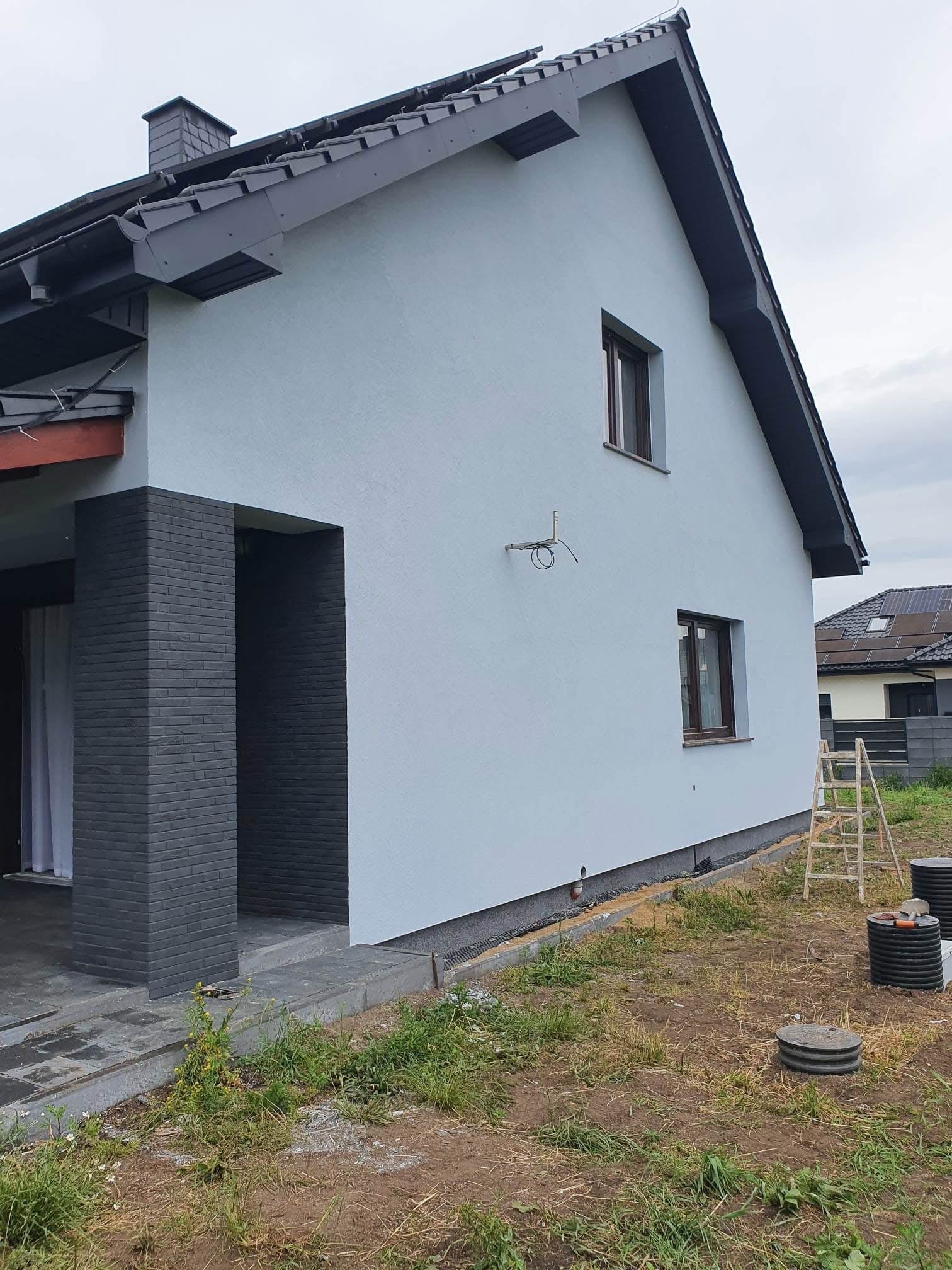 Jasnoszary dom jednorodzinny po renowacji elewacji, z ciemnoszarą stolarką okienną i wykończeniem z cegły klinkierowej przy wejściu. Widoczna drabina i teren wokół domu.