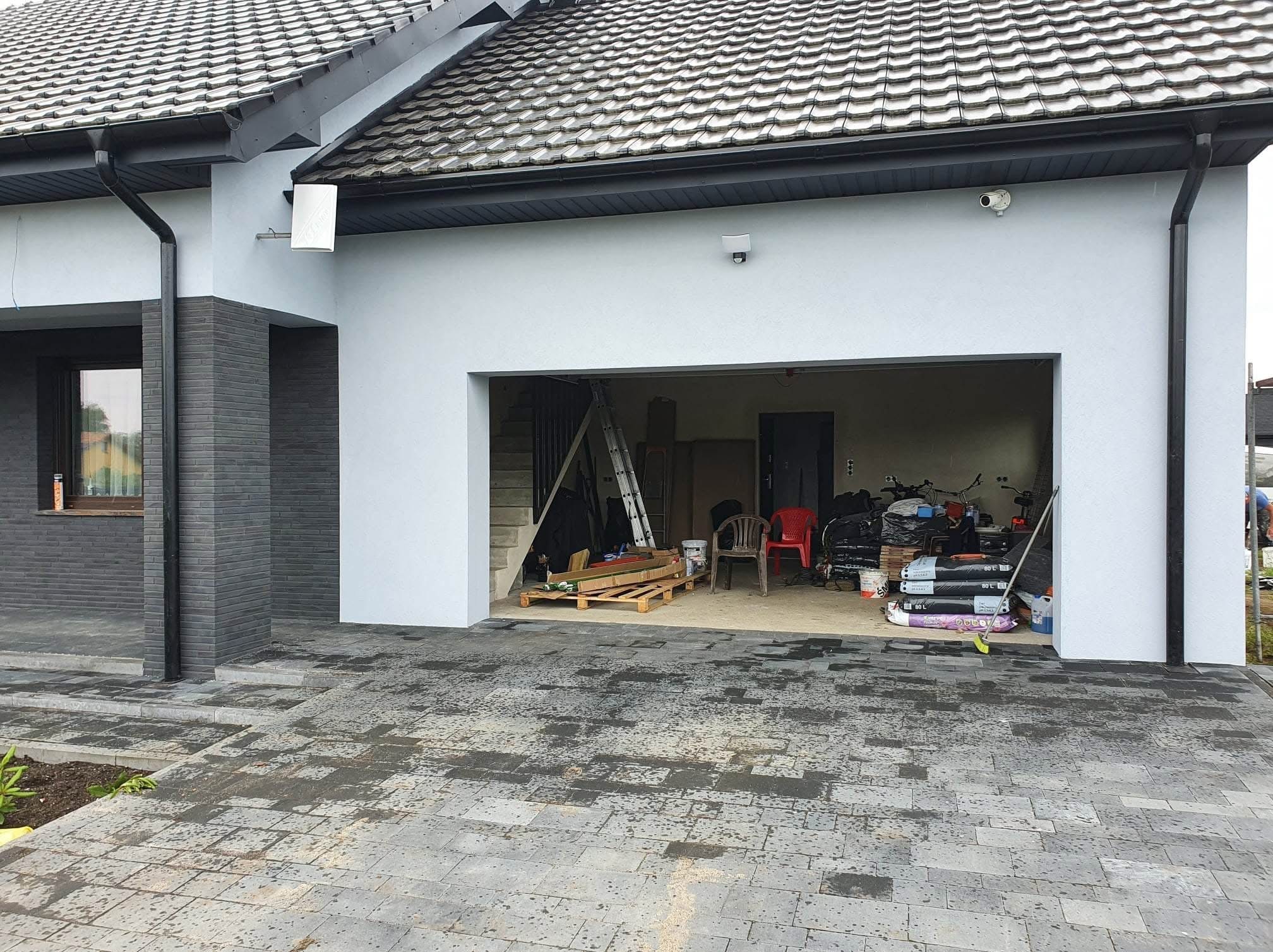 Nowoczesny dom jednorodzinny z jasną elewacją i grafitowymi elementami. Widoczny otwarty garaż z wyposażeniem i kostka brukowa na podjeździe.