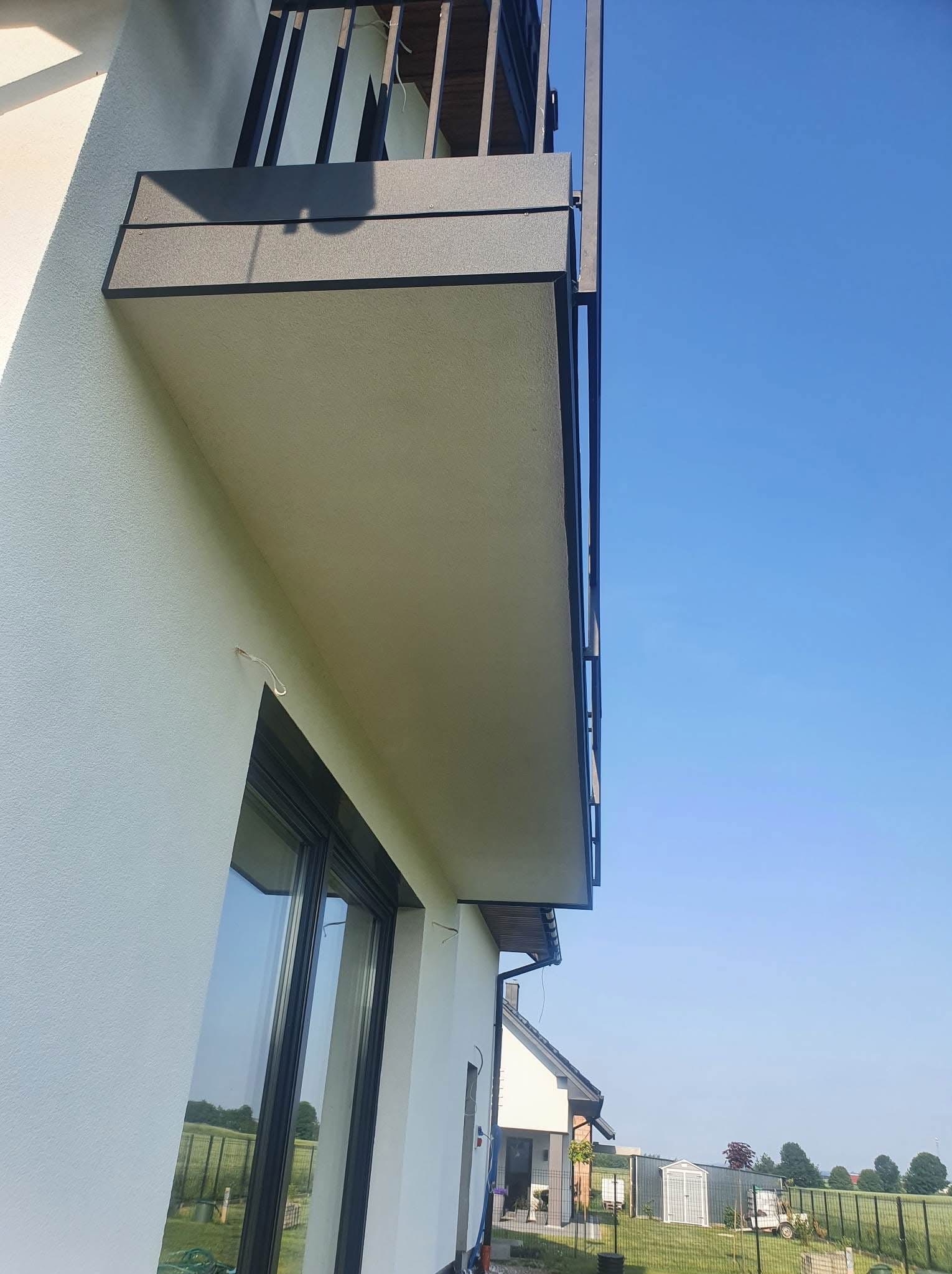 Boczny widok balkonu z czarną balustradą i elewacji domu w jasnym odcieniu, z widocznym oknem i fragmentem otoczenia.