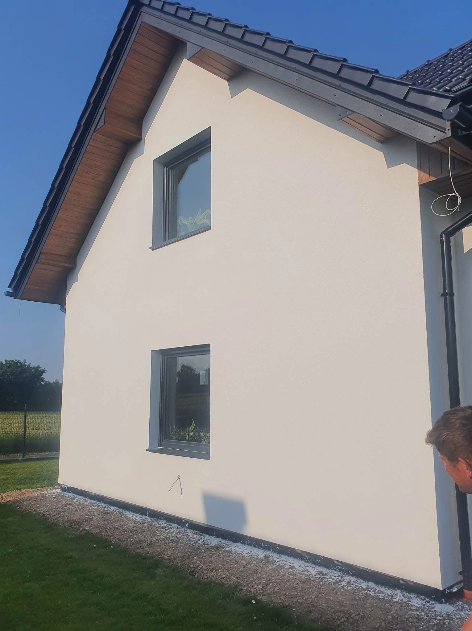 Boczny widok otynkowanego domu jednorodzinnego we Wrocławiu z dwoma oknami i ciemną dachówką. Elewacja w odcieniach bieli. Widoczne fragmenty ogrodu.