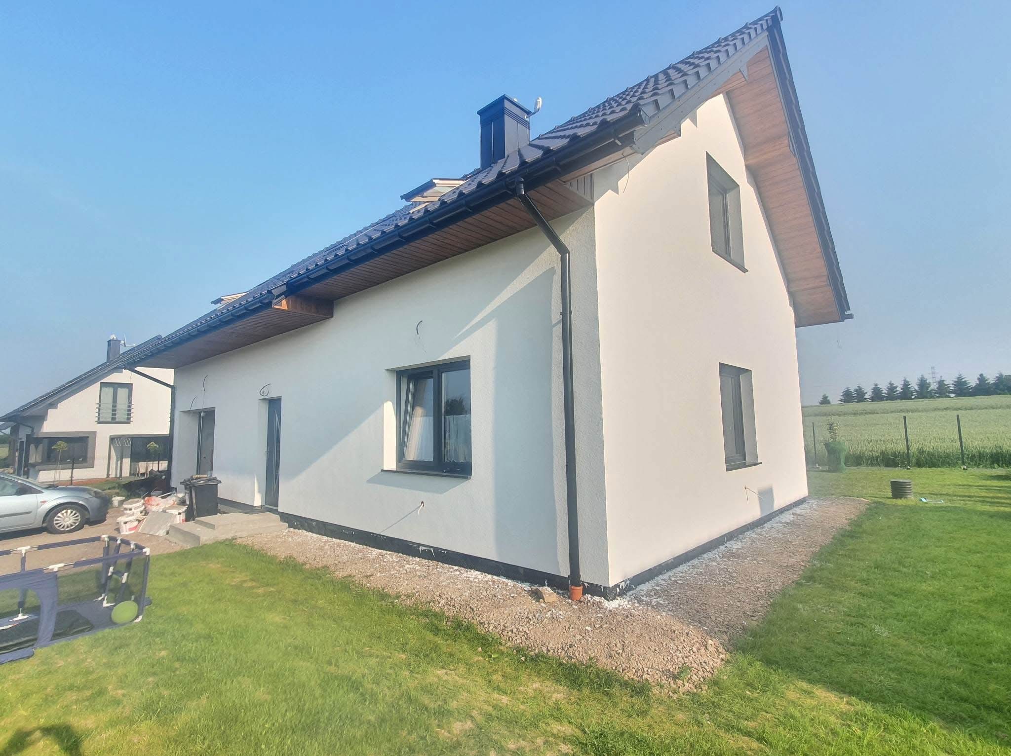 Biała elewacja domu jednorodzinnego z ciemną stolarką okienną i dachem. Widoczne ślady prac wykończeniowych wokół budynku. W tle zieleń i błękitne niebo.