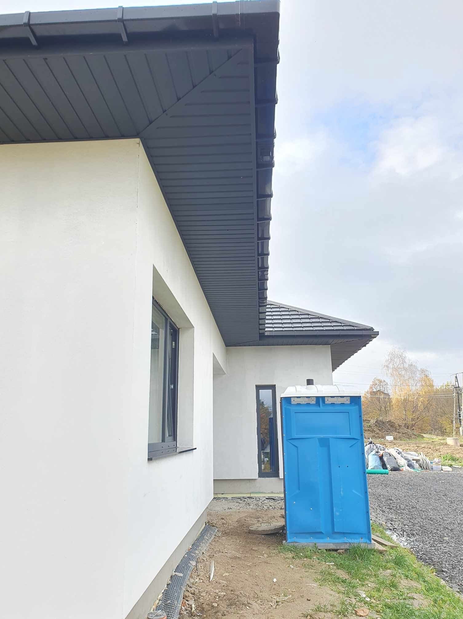 Boczny widok domu z białą elewacją i szarym dachem. Na pierwszym planie niebieska toaleta przenośna. Widoczne okno z czarną ramą i fragment krajobrazu w tle.