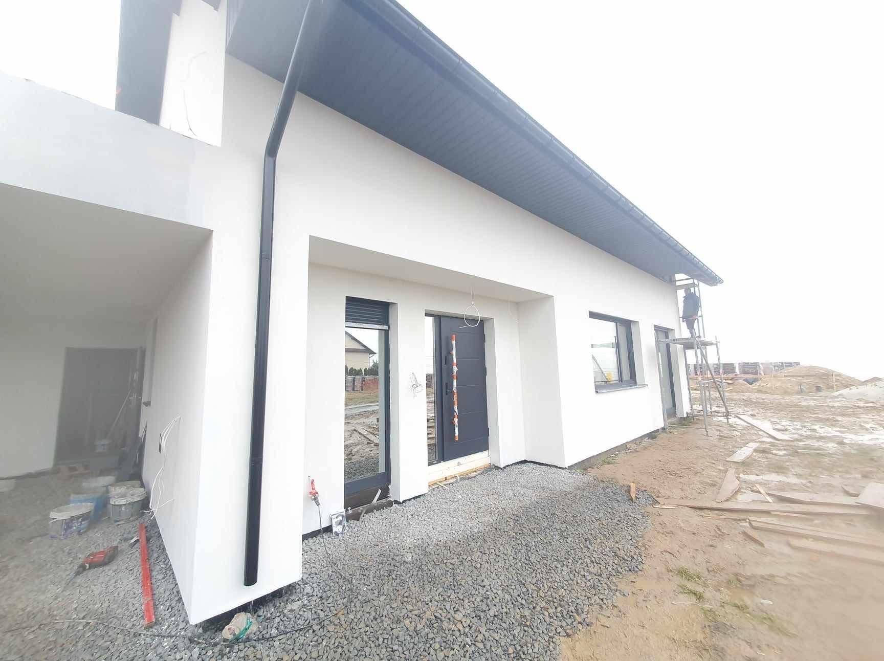 Nowoczesny dom jednorodzinny po malowaniu elewacji, z ciemnoszarymi oknami i dachem. Prace wykończeniowe na zewnątrz, widoczny pracownik na rusztowaniu.