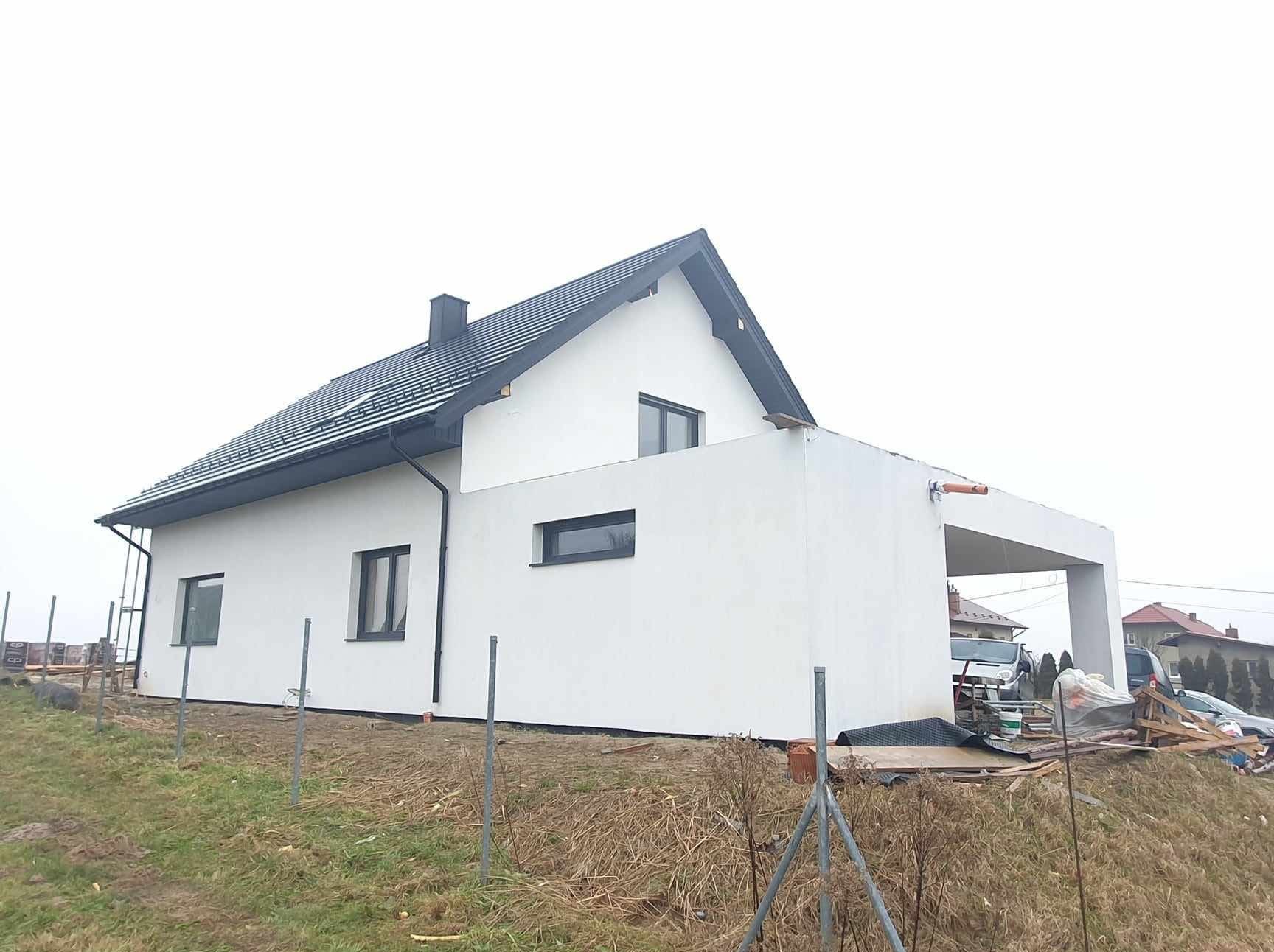 Nowoczesny dom z białą elewacją i ciemną stolarką okienną, garaż w bryle budynku, widok z zewnątrz na tle pochmurnego nieba. Elementy wykończeniowe w toku.