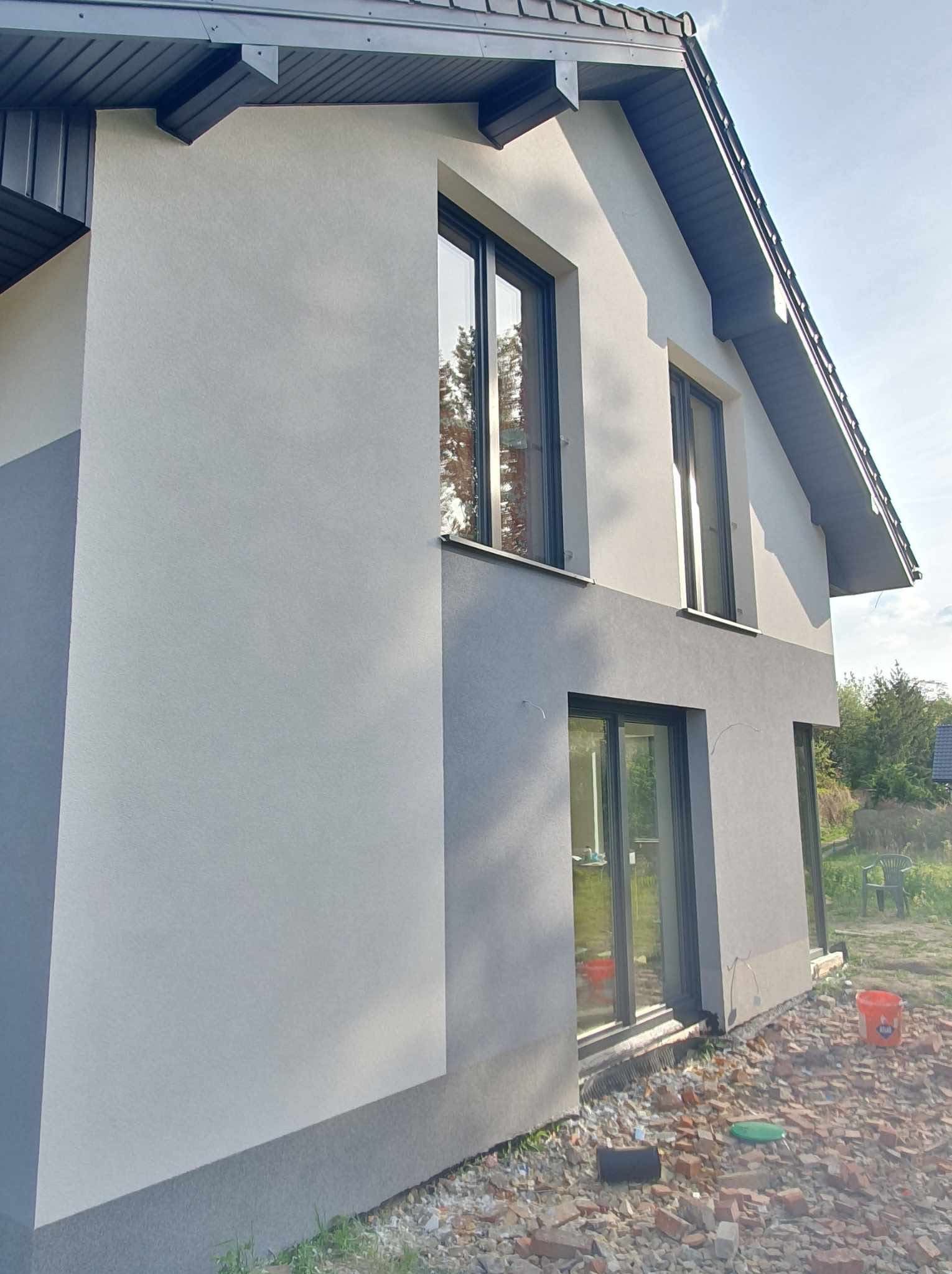 Dwukolorowa elewacja domu jednorodzinnego z ciemnymi oknami, widok z boku, z elementami ogrodu w tle. Na ziemi gruz i wiadro.