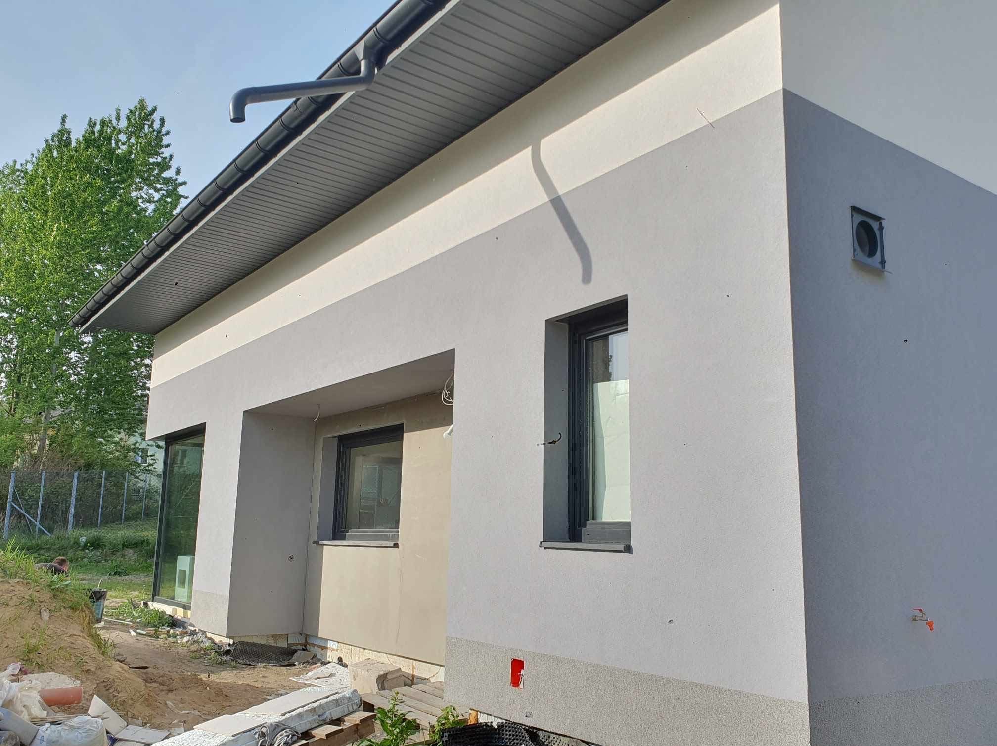 Dwukolorowa elewacja domu jednorodzinnego w odcieniach szarości, z widocznymi oknami w ciemnych ramach i fragmentem dachu z rynną. Budynek w trakcie wykończenia.