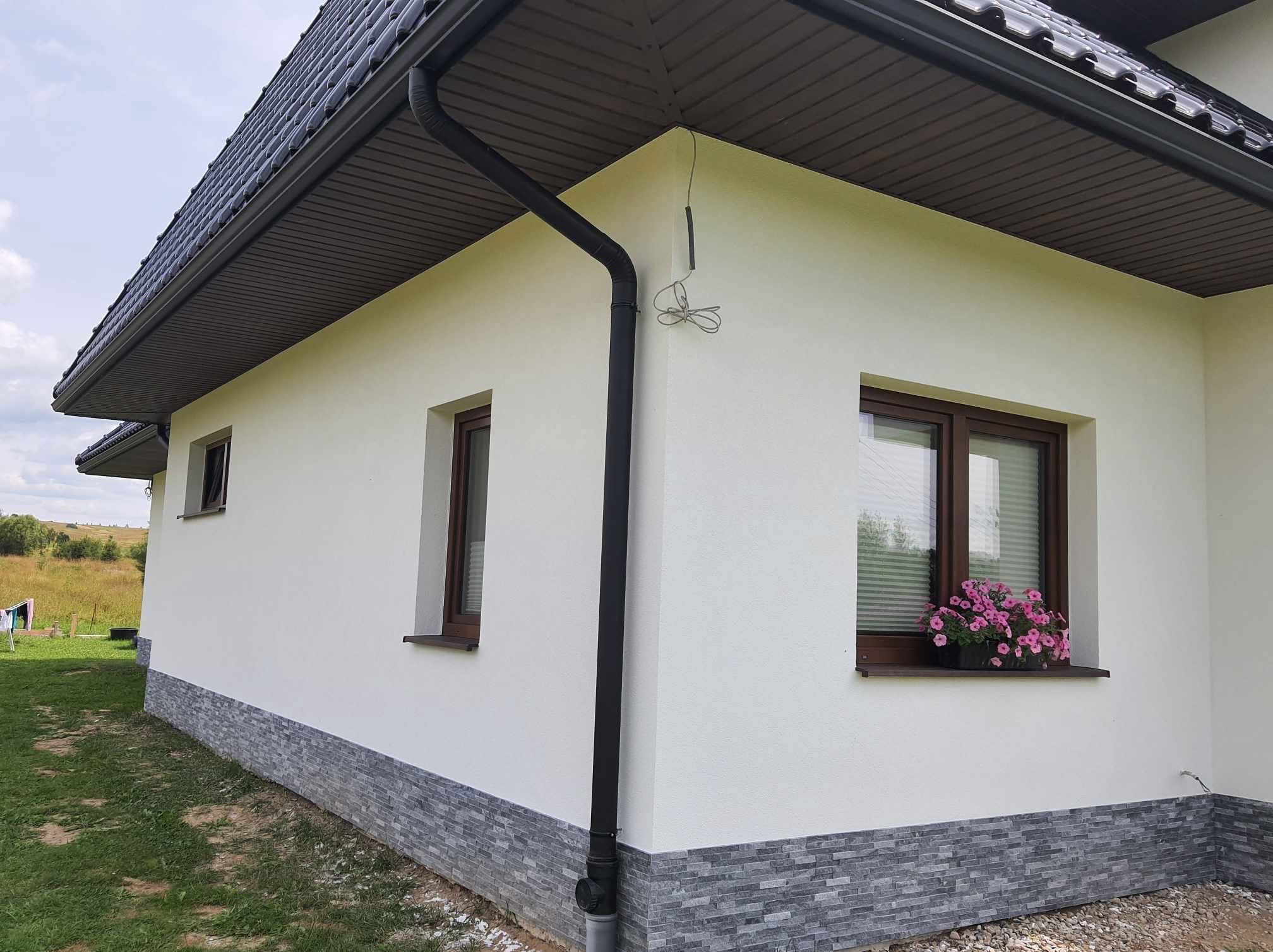Biała elewacja domu z brązowymi oknami i ciemnym dachem. W oknie różowe kwiaty, a dół elewacji wykończony szarą płytką imitującą kamień. Widoczny czarny system rynnowy.