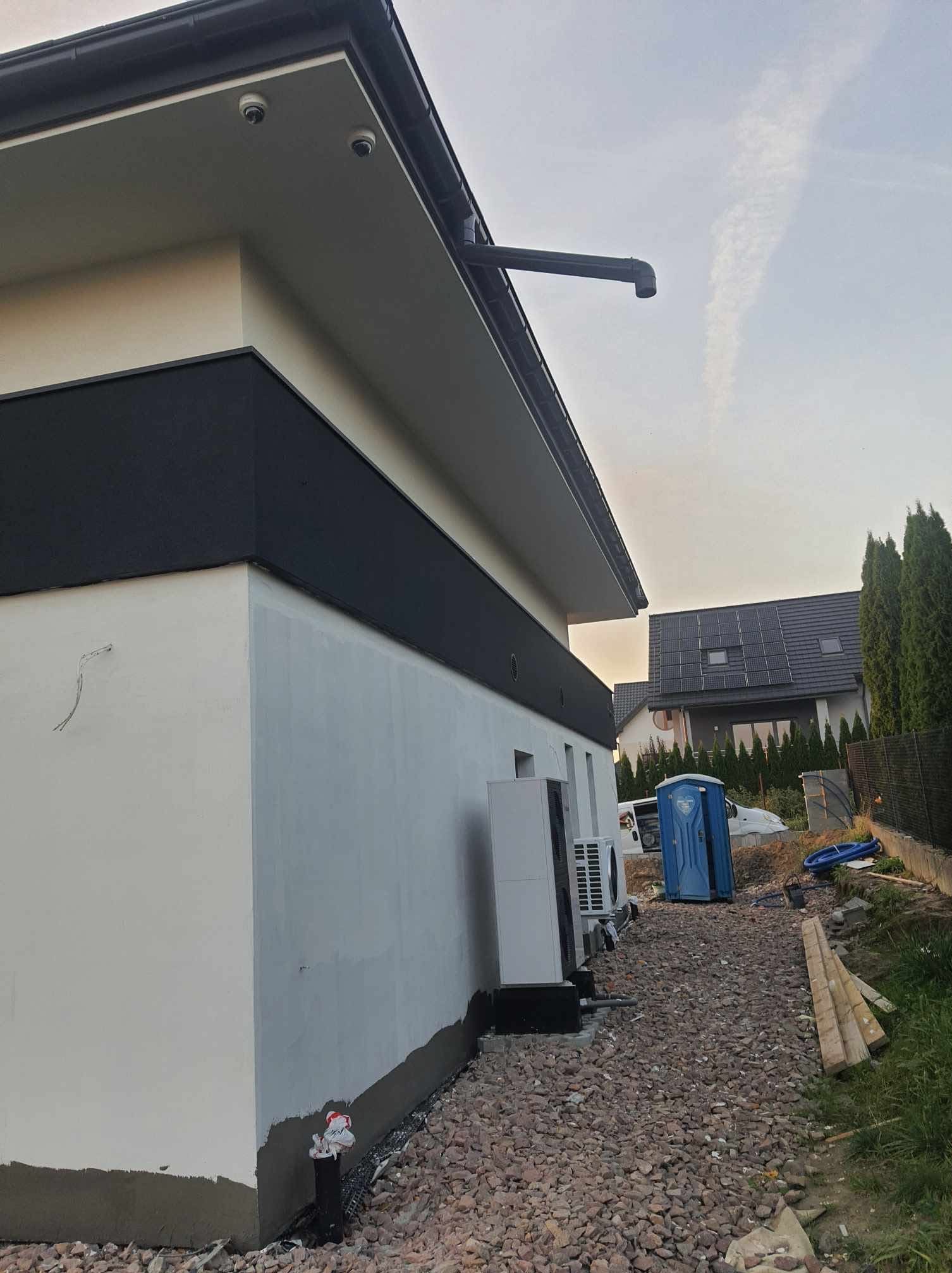 Boczny widok domu z zainstalowaną pompą ciepła i wykończoną elewacją w kolorze białym i czarnym. Widoczne kamery na dachu i panele słoneczne na sąsiednim budynku.