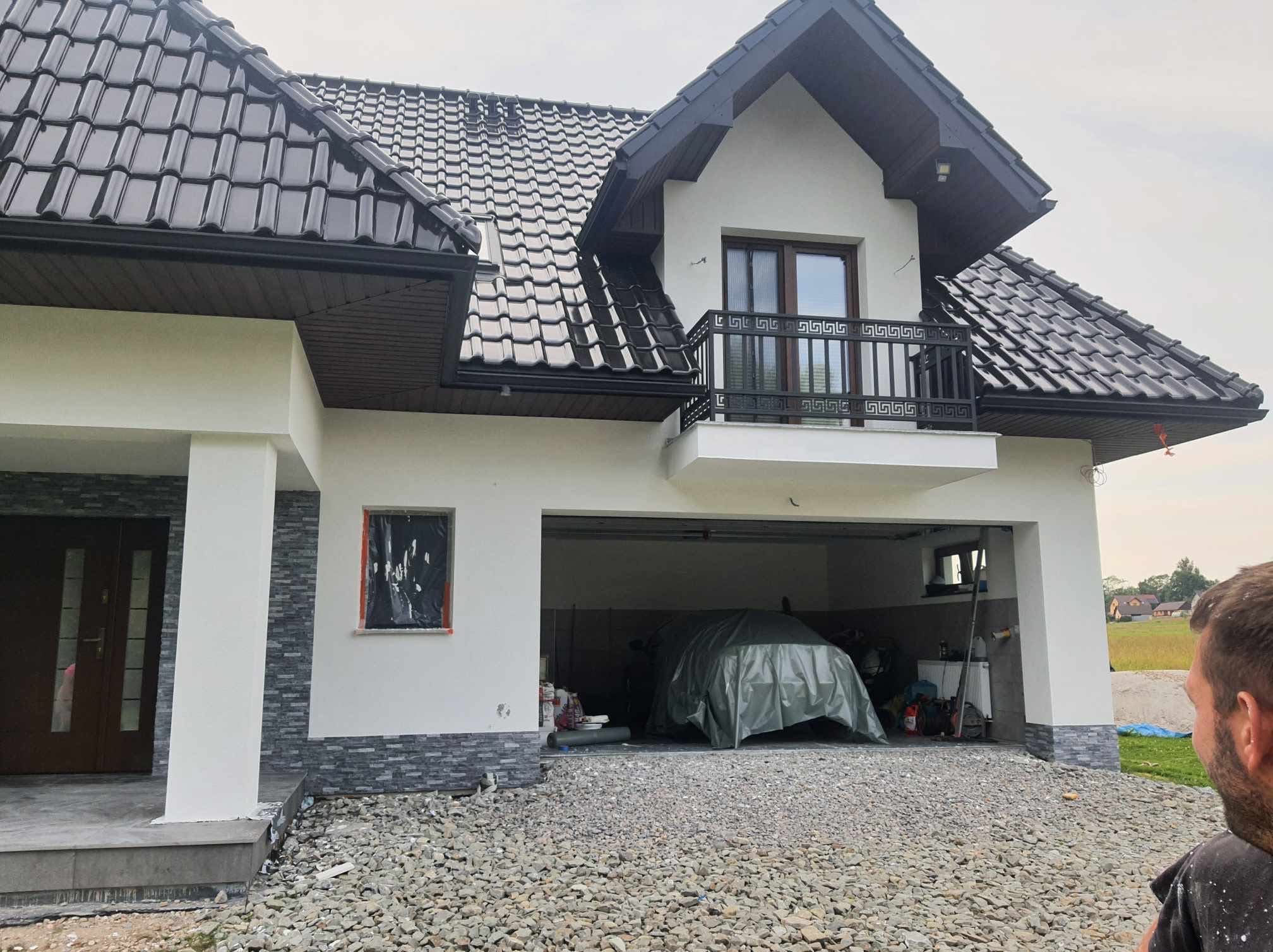 Dom z ciemną dachówką i jasną elewacją, balkon z ozdobną balustradą. Okno zabezpieczone folią, garaż otwarty z przykrytym samochodem. Podłoże żwirowe.