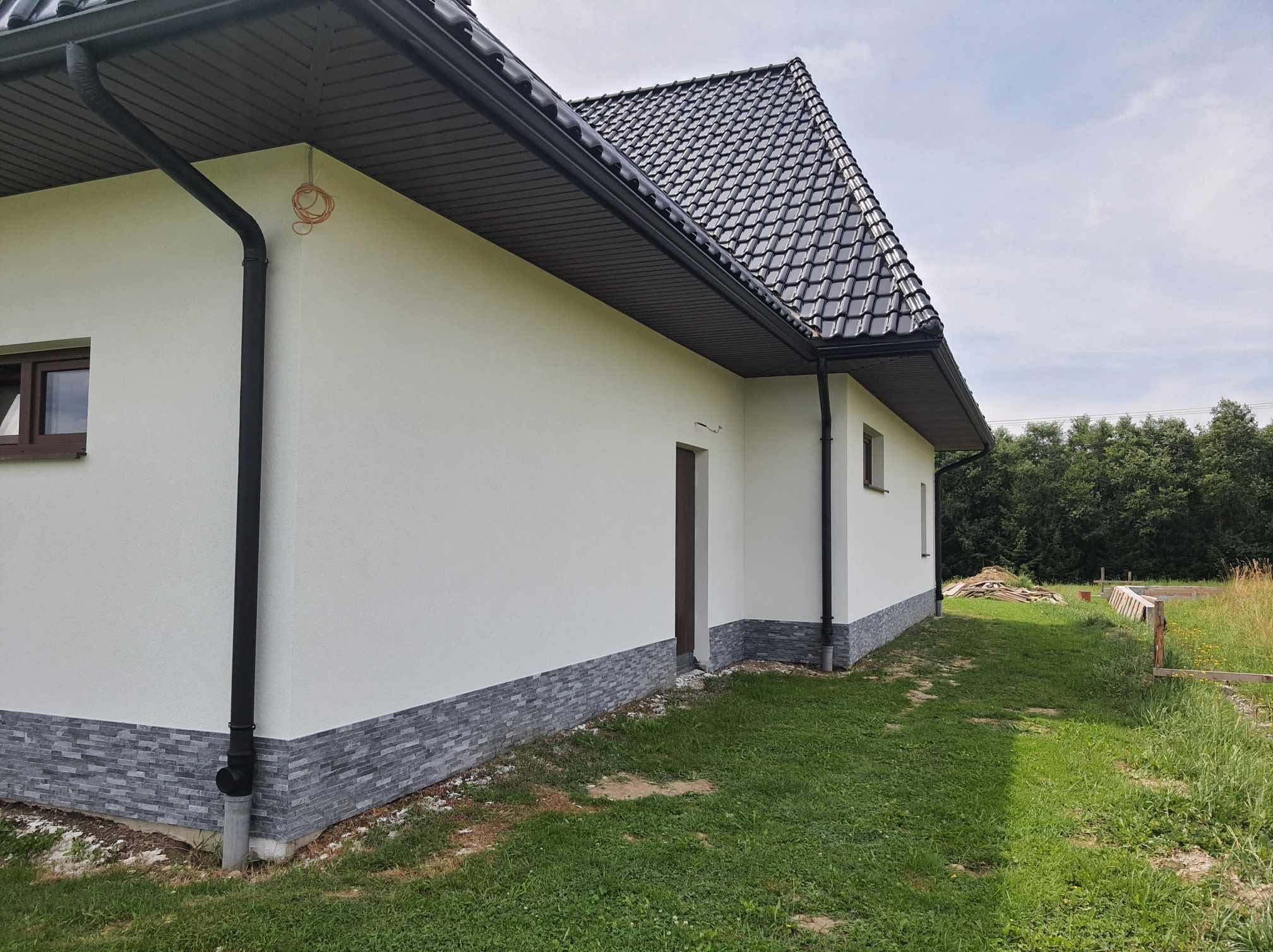 Wykończona elewacja domu jednorodzinnego z szarą podmurówką imitującą kamień i białym tynkiem. Widoczny fragment dachu z czarną dachówką i rynną, oraz fragment ogrodu.