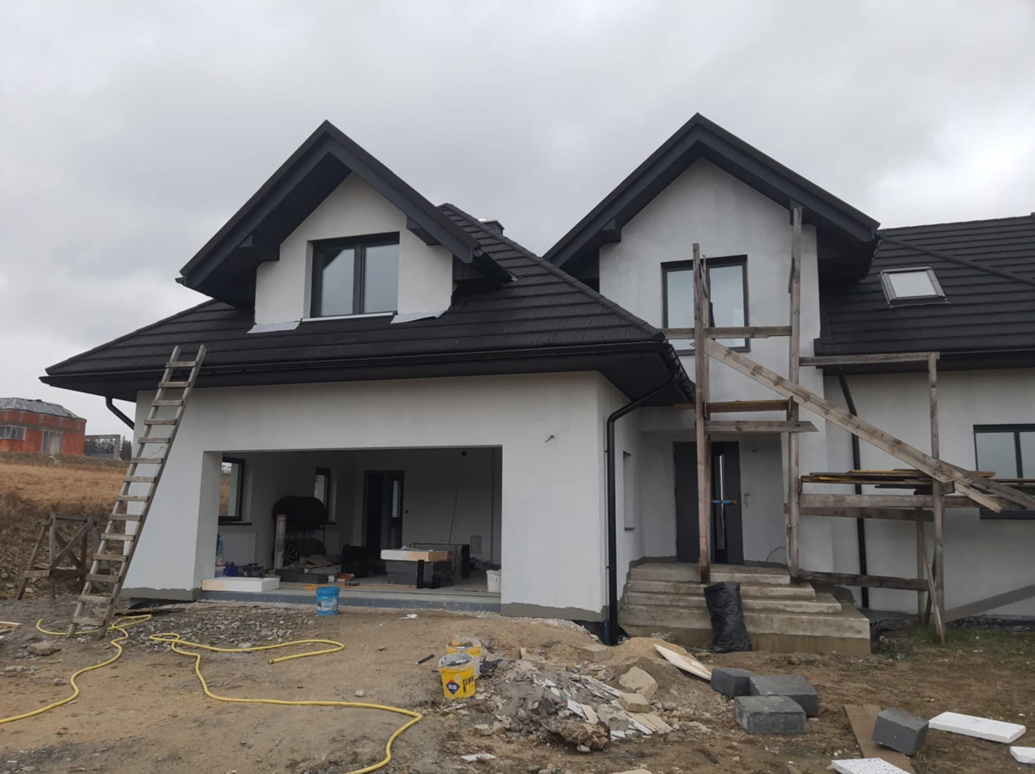 Dom jednorodzinny w trakcie prac elewacyjnych. Widoczne rusztowania, drabina oparta o ścianę, teren wokół domu z materiałami budowlanymi i narzędziami malarskimi.