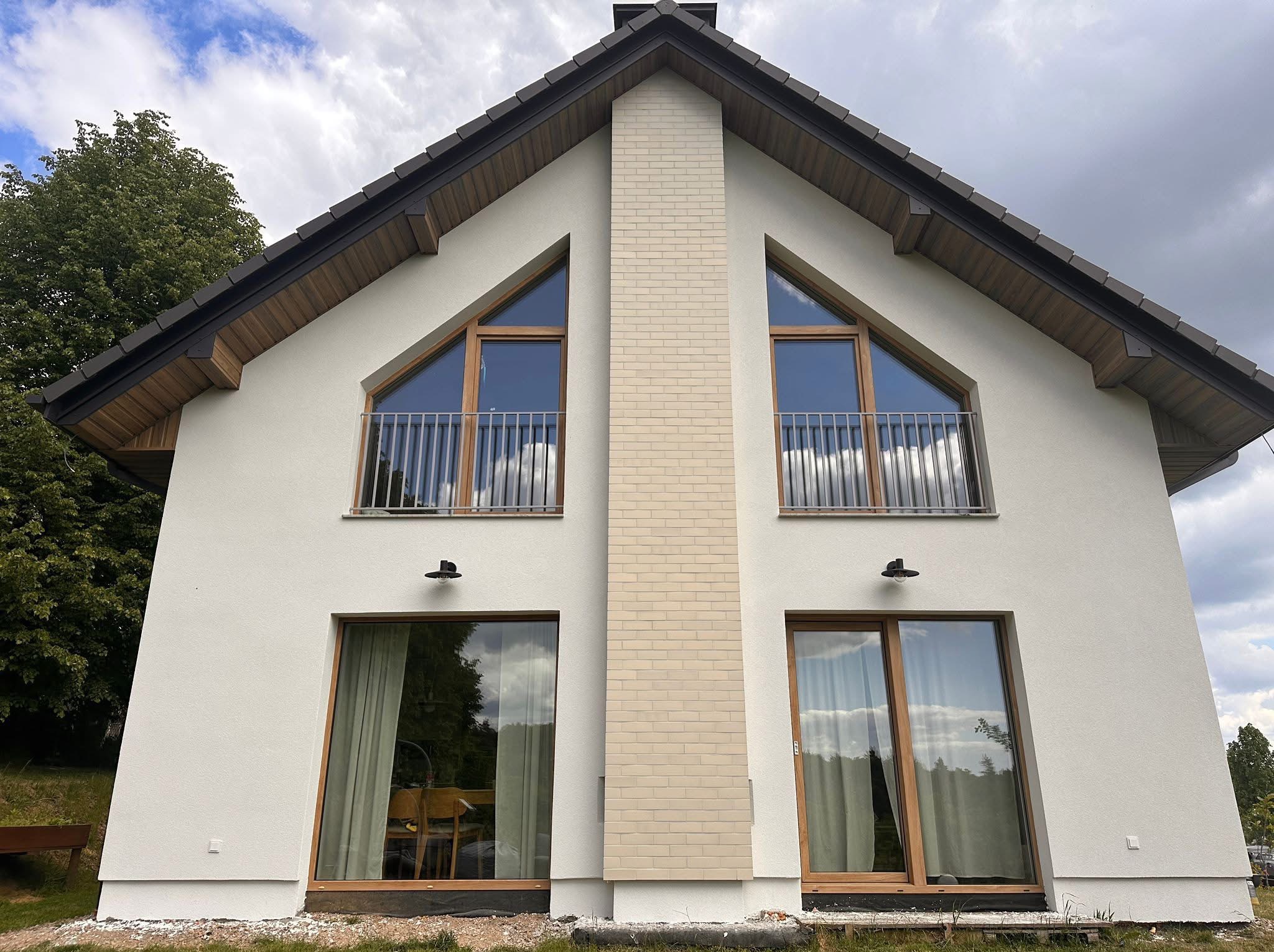 Nowoczesny dom z białą elewacją, drewnianymi oknami i ceglanym kominem, uchwycony w pochmurny dzień z niskiego kąta.
