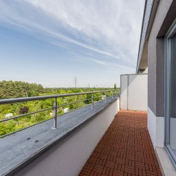 Freedom - Przemysław Baran - Przestronny balkon z brązową podłogą, metalową barierką i widokiem na zieleń oraz błękitne niebo z delikatnymi chmurami. Nowoczesne wykończenie i jasne otoczenie.