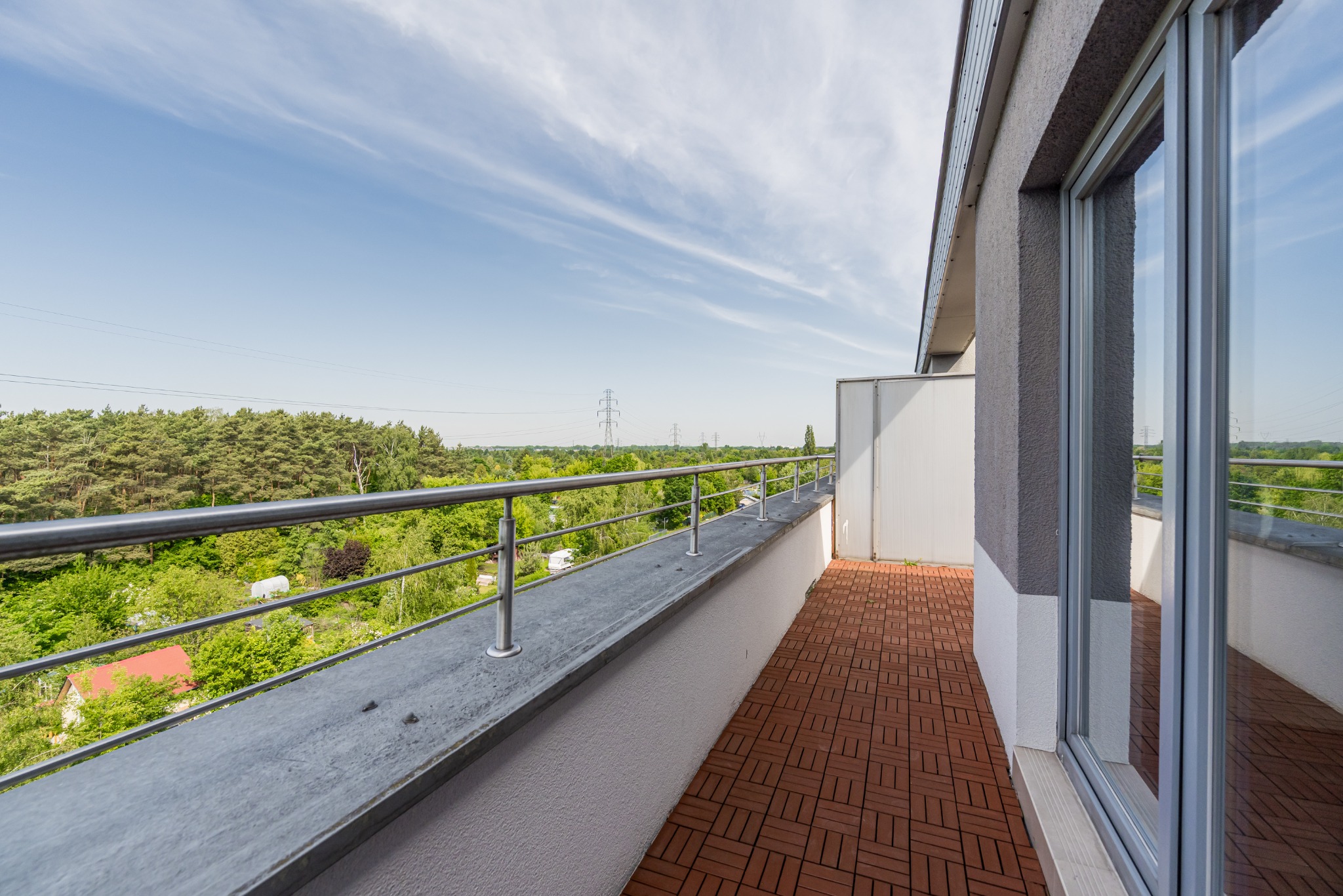 Przestronny balkon z brązową podłogą, metalową barierką i widokiem na zieleń oraz błękitne niebo z delikatnymi chmurami. Nowoczesne wykończenie i jasne otoczenie.