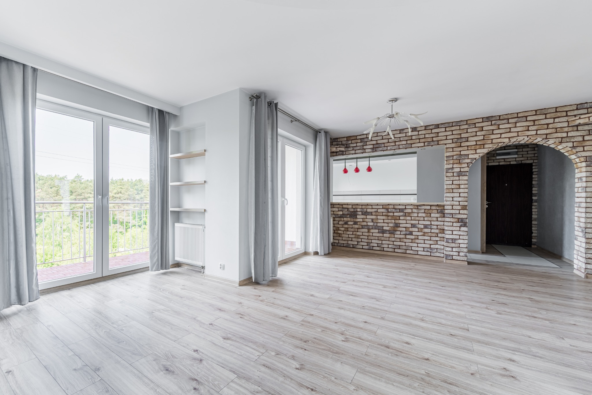 Jasne, przestronne wnętrze po remoncie w Markach: parkiet, ceglana ściana, okna balkonowe z widokiem na zieleń. Nowoczesne oświetlenie i minimalistyczne półki.