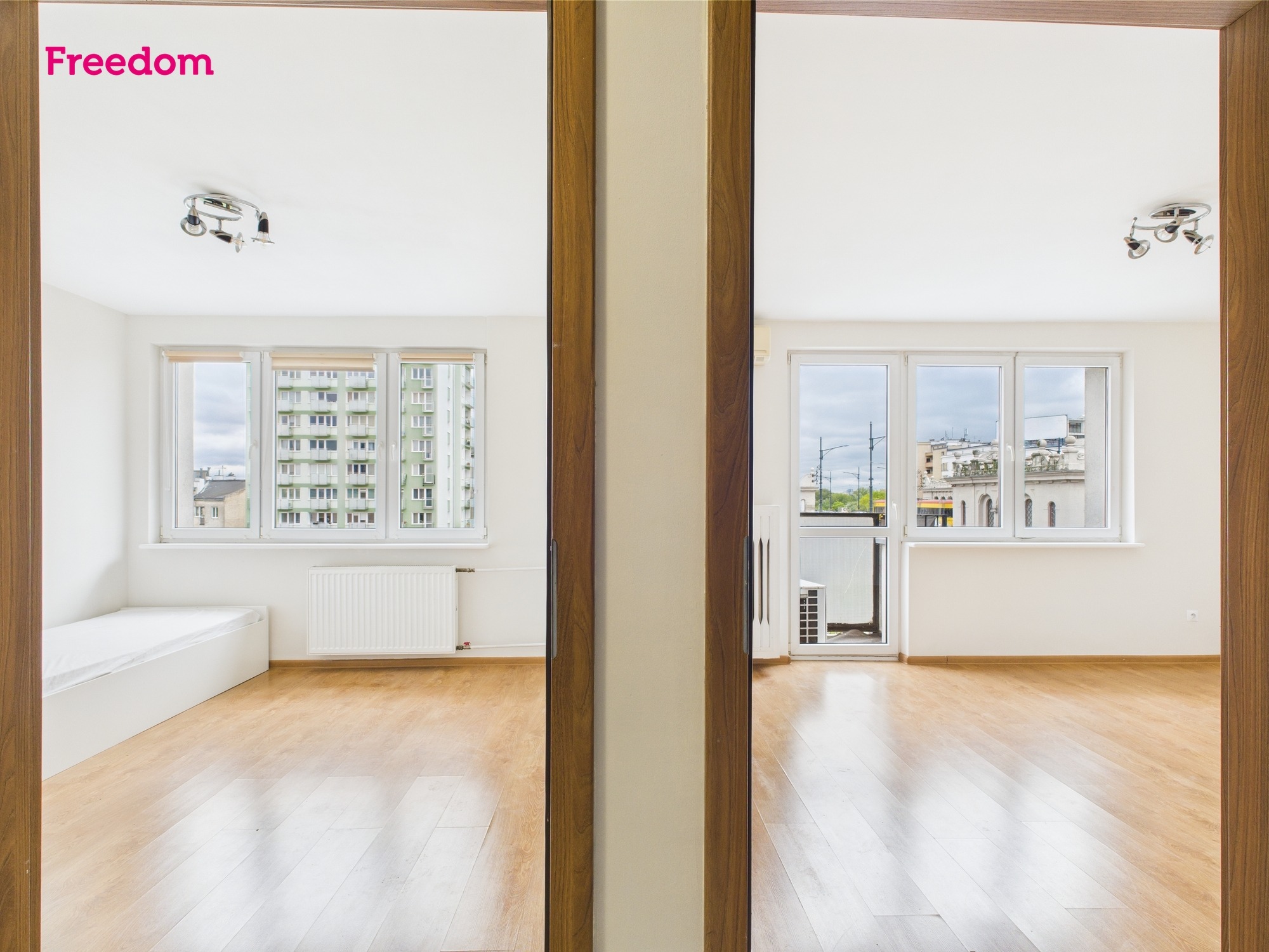 Jasne, puste pokoje z drewnianą podłogą i oknami z widokiem na miasto, oddzielone ścianą z drewnianymi elementami. Minimalistyczny wystrój, dużo światła dziennego.