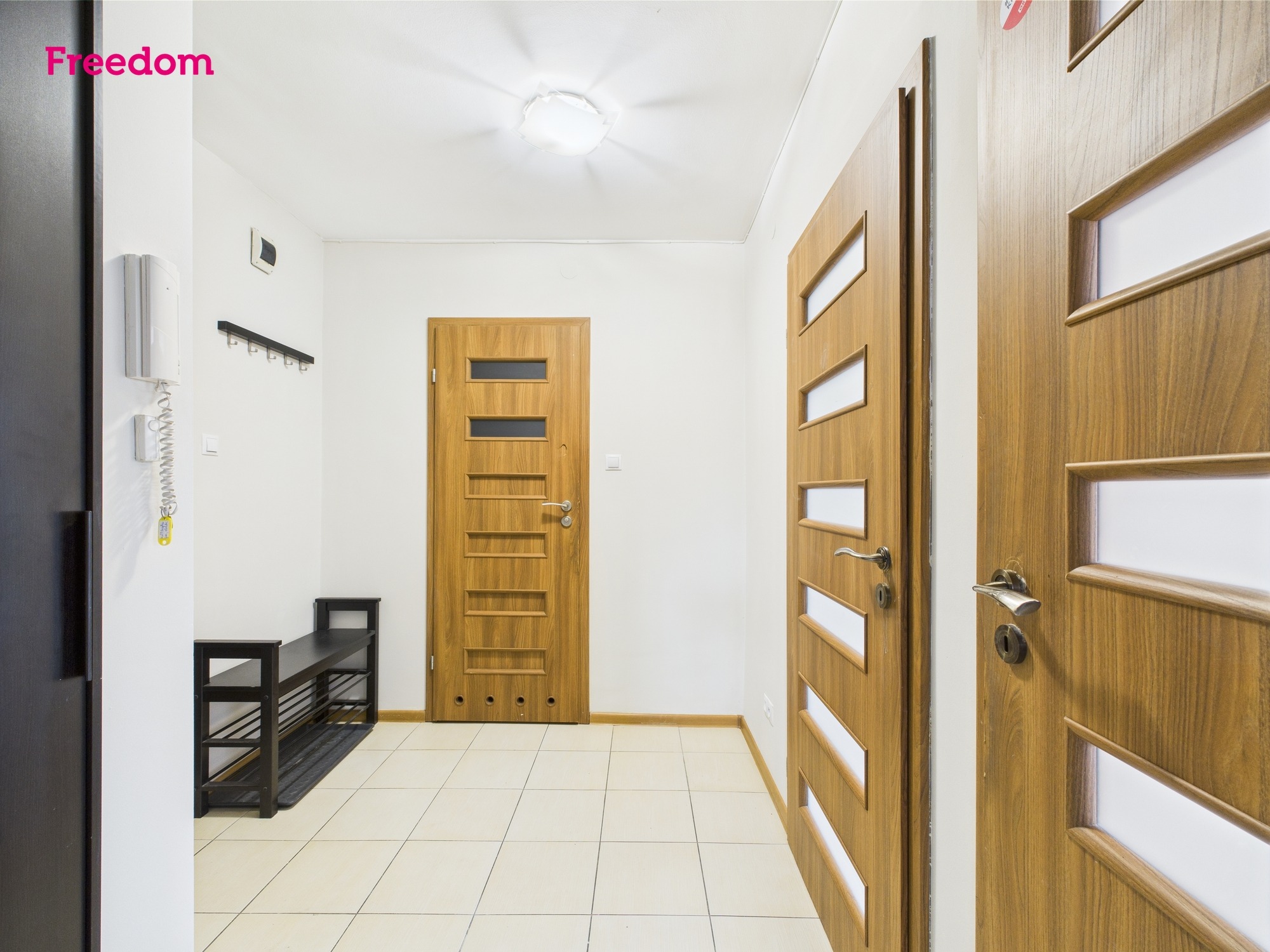 Jasny korytarz z drewnianymi drzwiami z przeszkleniami, białe ściany i podłoga w jasnych płytkach. W tle widać wieszak i czarną ławkę na buty.