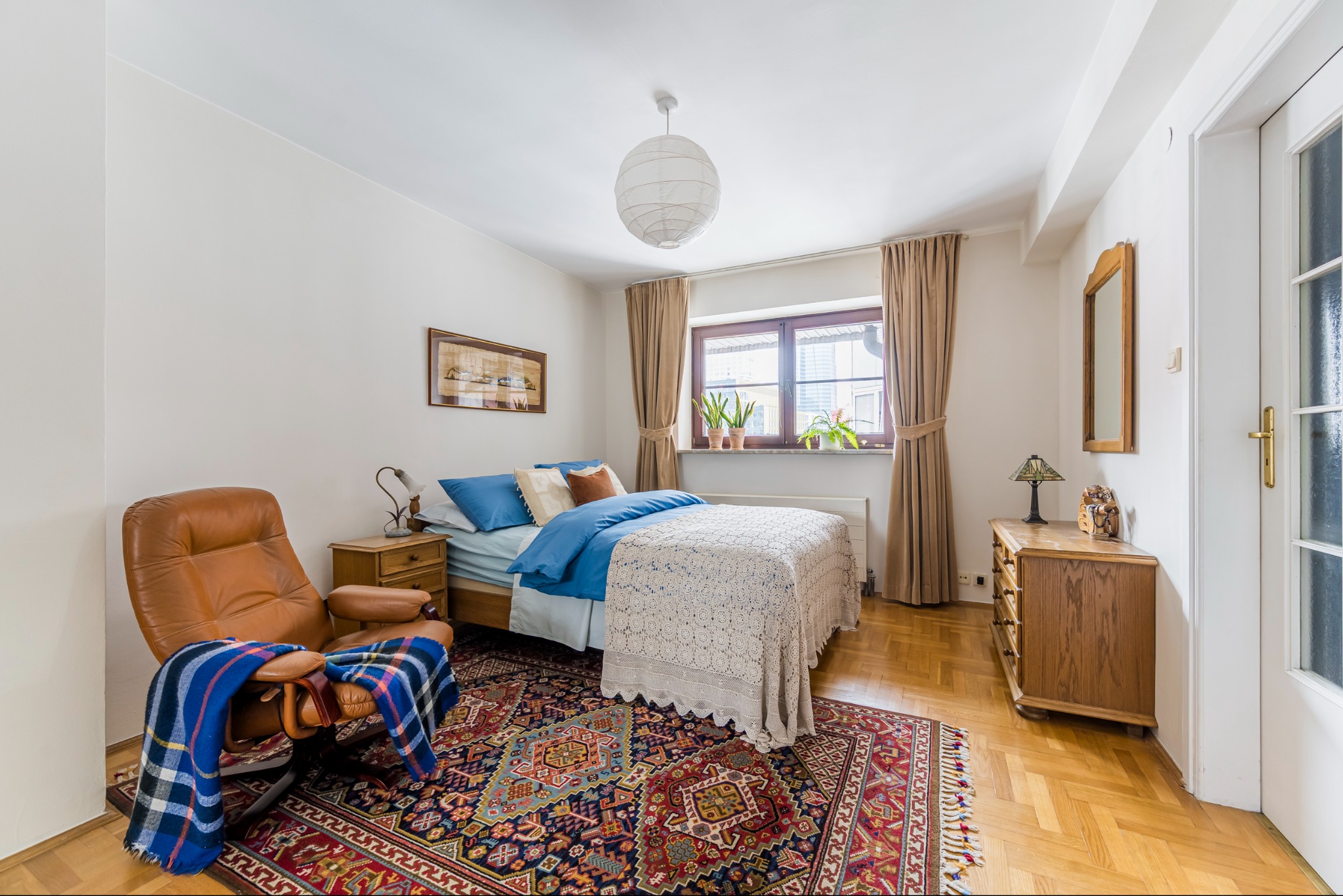 Przytulna sypialnia z drewnianą komodą, brązowym fotelem i wzorzystym dywanem. Na parapecie doniczki z roślinami, a na ścianie obrazek.