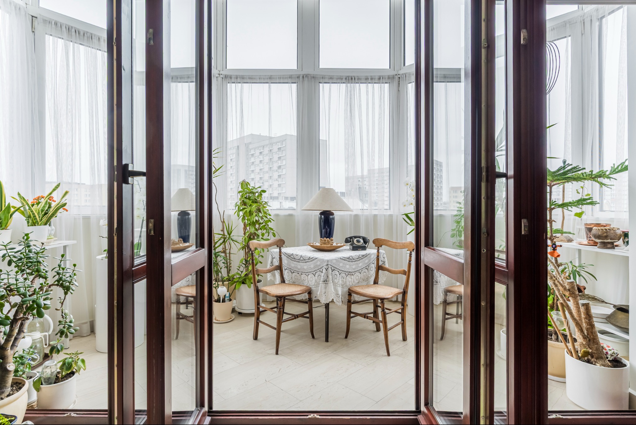 Otwarty balkon z roślinami w donicach, stolikiem nakrytym koronkowym obrusem i krzesłami. W tle budynki mieszkalne za firankami.