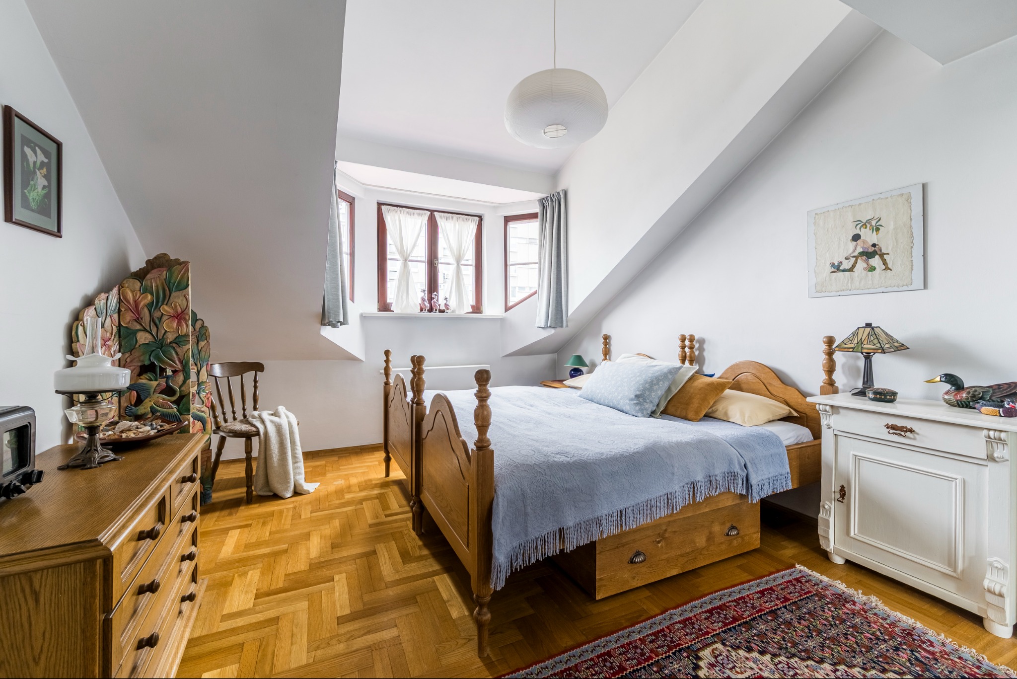 Przytulna sypialnia na poddaszu z drewnianym łóżkiem, komodą i parkietem w jodełkę. Okno z firankami w skosie dachu. Dekoracje w stylu vintage, ciepłe oświetlenie.