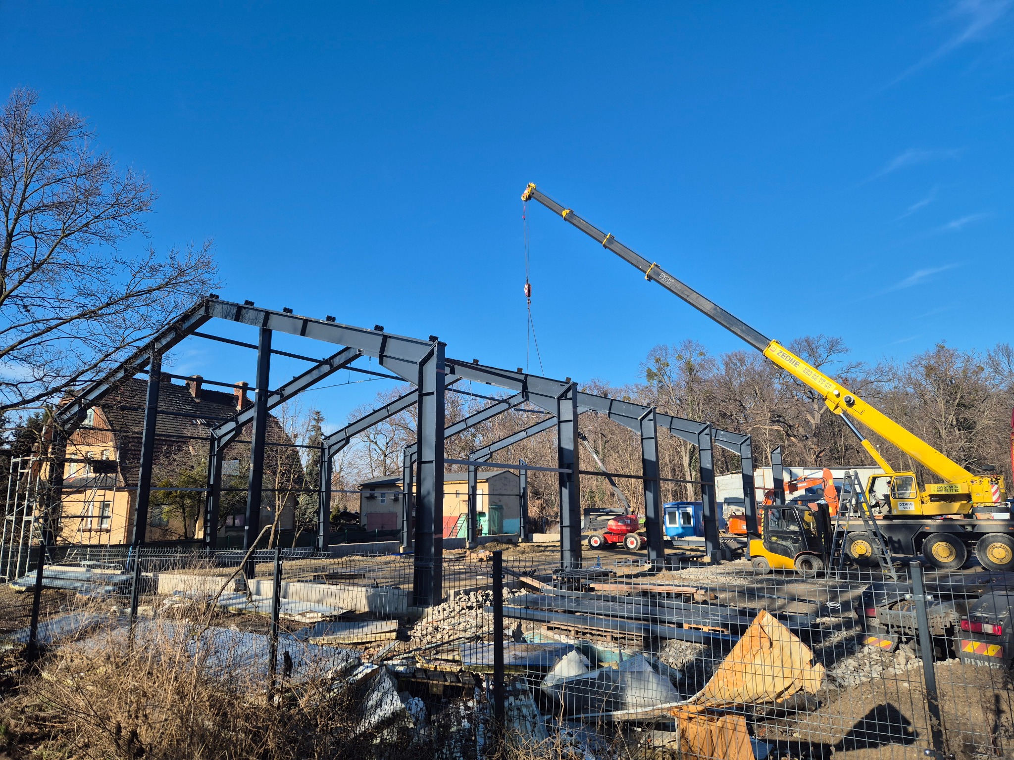 Stalowa konstrukcja hali w trakcie montażu z użyciem żółtego dźwigu na tle błękitnego nieba i zabudowań. Widoczne maszyny budowlane i ogrodzenie placu budowy.