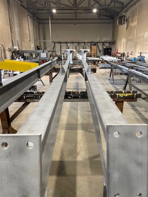 Dwie równoległe, szare, metalowe belki z otworami, leżące w warsztacie. W tle widoczne inne konstrukcje i narzędzia w przestrzeni roboczej.