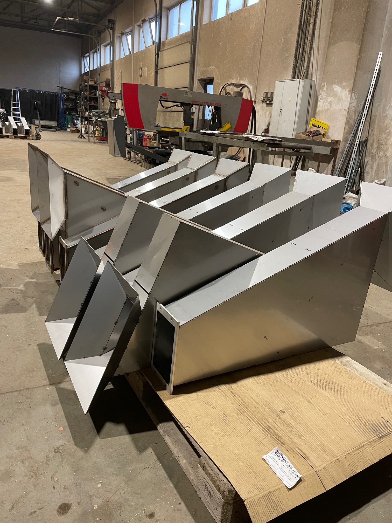 Szereg gotowych, spawanych, stalowych elementów wentylacyjnych na palecie w hali produkcyjnej. W tle widoczna piła taśmowa do metalu.