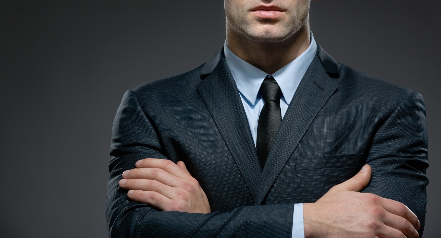 Mężczyzna w granatowym garniturze i czarnym krawacie, z założonymi rękami, prezentuje profesjonalny wizerunek na tle szarego studia.