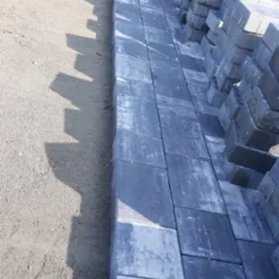 Fragment układanej szarej kostki brukowej na podsypce piaskowej, z ułożonymi obok stosami kostki. Widoczne narzędzie i taczka w tle. Ujęcie z góry, w słoneczny dzień.