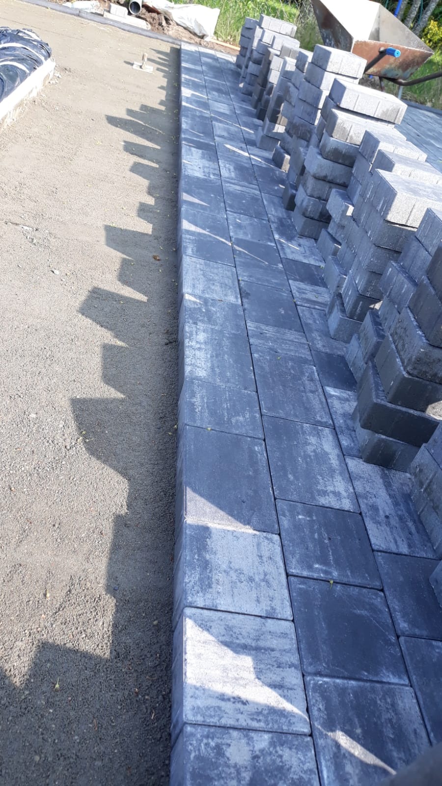 Fragment układanej szarej kostki brukowej na podsypce piaskowej, z ułożonymi obok stosami kostki. Widoczne narzędzie i taczka w tle. Ujęcie z góry, w słoneczny dzień.