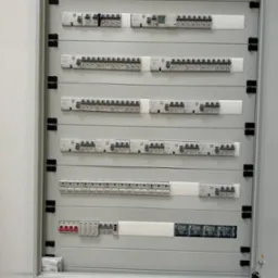 Rozdzielnica elektryczna z bezpiecznikami, szyny DIN i okablowanie wewnątrz szafy. Widoczne elementy instalacji elektrycznej, szare tło, oświetlenie sufitowe.